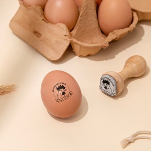 Puede incluir: Un huevo marr&oacute;n con un sello de goma negro y blanco que dice "Rosemary Farm, Fresh with Love" y un sello de goma de madera con un dise&ntilde;o de gallo.