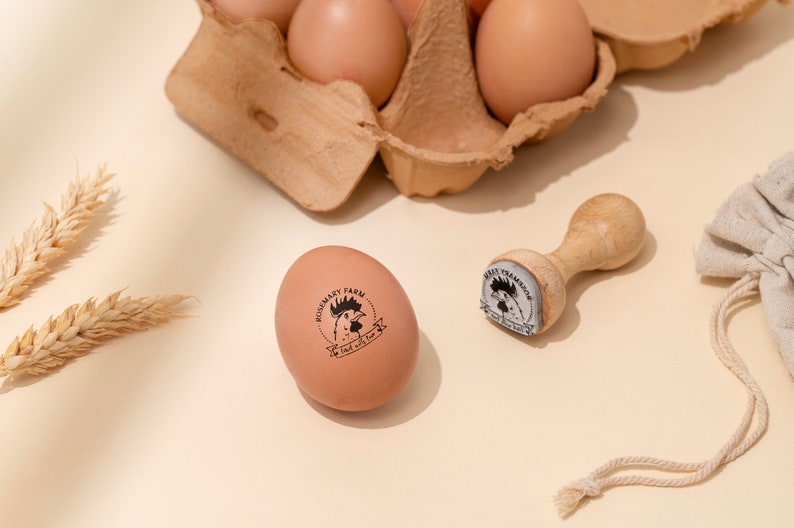 Puede incluir: Un huevo marr&oacute;n con un sello de goma negro y blanco que dice "Rosemary Farm, Fresh with Love" y un sello de goma de madera con un dise&ntilde;o de gallo.