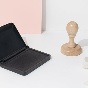 Op de afbeelding: Een zwart inktkussen met het deksel open, naast een houten stempel. Het inktkussen is rechthoekig en de stempel heeft een ronde basis en een afgeronde handgreep. Er is ook een wit lint aanwezig.