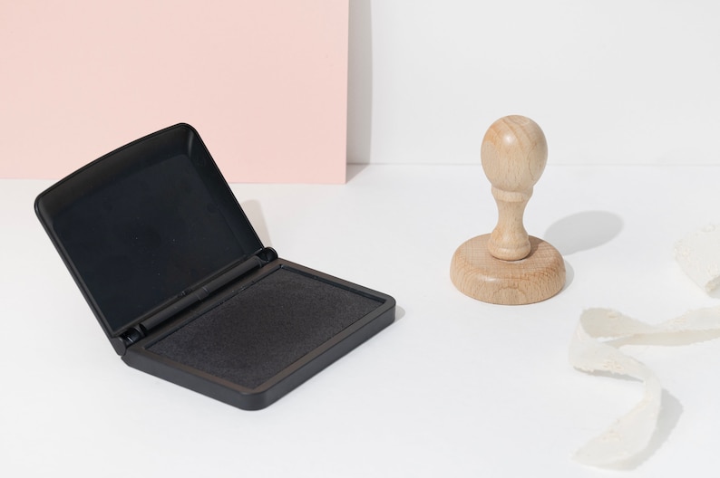 Op de afbeelding: Een zwart inktkussen met het deksel open, naast een houten stempel. Het inktkussen is rechthoekig en de stempel heeft een ronde basis en een afgeronde handgreep. Er is ook een wit lint aanwezig.