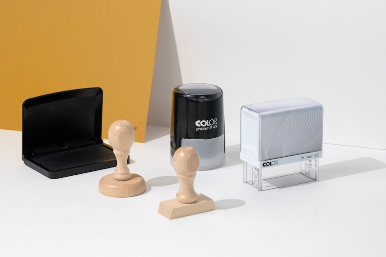 May include: An assortment of office stamps and an ink pad. The stamps include a wooden stamp with a round handle, a rectangular wooden stamp, a black and gray stamp, and a clear stamp with the text "Printer 40".