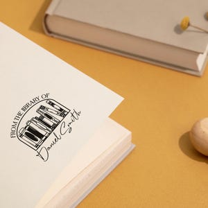 May include: A white book with a black stamp that reads "From the library of Daniel Smith" with a graphic of a bookshelf with books. The stamp is on the front cover of the book.