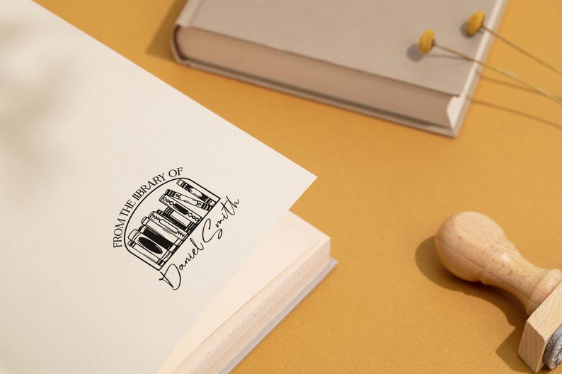May include: A white book with a black stamp that reads "From the library of Daniel Smith" with a graphic of a bookshelf with books. The stamp is on the front cover of the book.