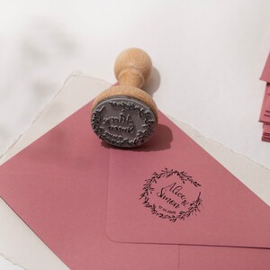 Op de afbeelding: Een houten stempel met een cirkelvormig ontwerp, rustend op een stoffige roze envelop. De stempel heeft een houten handvat en een rubberen stempel met de woorden "Alice & Simon" en de datum. Een stapel bijpassende enveloppen staat op de achtergrond.
