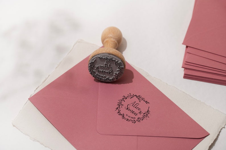 Op de afbeelding: Een houten stempel met een cirkelvormig ontwerp, rustend op een stoffige roze envelop. De stempel heeft een houten handvat en een rubberen stempel met de woorden "Alice & Simon" en de datum. Een stapel bijpassende enveloppen staat op de achtergrond.