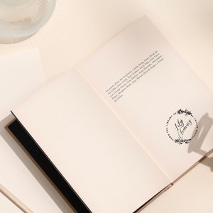 May include: Open book with black binding and cream-colored pages. The text "THE LIBRARY OF JULY CLEMENCY" is printed inside a floral wreath. A pencil, a gold paper clip, and a glass bowl are on the off-white surface.