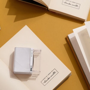 May include: An open book with blank pages, a white book with the text "FROM THE LIBRARY OF - Blandon Smith", and a white and clear stamp with the text "Printer 40" and "COLOR" on a mustard-colored surface. The image suggests a stationery or book-related theme.