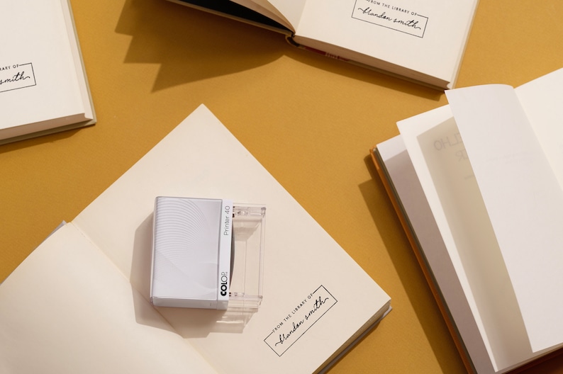 May include: An open book with blank pages, a white book with the text "FROM THE LIBRARY OF - Blandon Smith", and a white and clear stamp with the text "Printer 40" and "COLOR" on a mustard-colored surface. The image suggests a stationery or book-related theme.