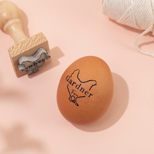 Puede incluir: Un huevo marr&oacute;n con un sello negro que dice "Gardner Farm" y una silueta de pollo. Tambi&eacute;n hay un sello de madera con una silueta de pollo en la imagen.