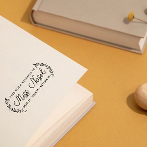 May include: A wooden stamp with the text "This book belongs to Miss Nosek, Read it, Love it, Return it" in a decorative frame, stamped on a book page. A book and dried flowers are in the background.