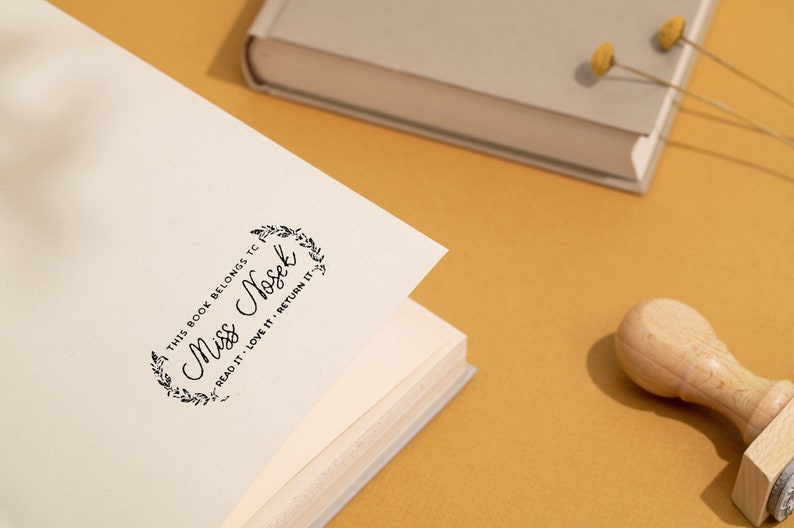 May include: A wooden stamp with the text "This book belongs to Miss Nosek, Read it, Love it, Return it" in a decorative frame, stamped on a book page. A book and dried flowers are in the background.
