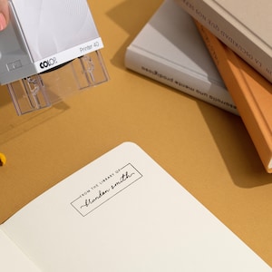 May include: A white and gray "Printer 40" stamp is being used to imprint text on a white notebook page. The text reads "FROM THE LIBRARY OF Blandan Smith". Several books with tan and gray covers are in the background.