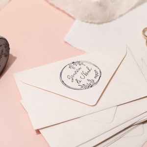 Op de afbeelding: Een houten stempel met een bloemenontwerp en de namen "Joanna & Noah" en een datum, naast een stapel cr&egrave;mekleurige enveloppen. Twee gouden trouwringen zijn zichtbaar op de achtergrond. De stempel staat op een lichtroze ondergrond.