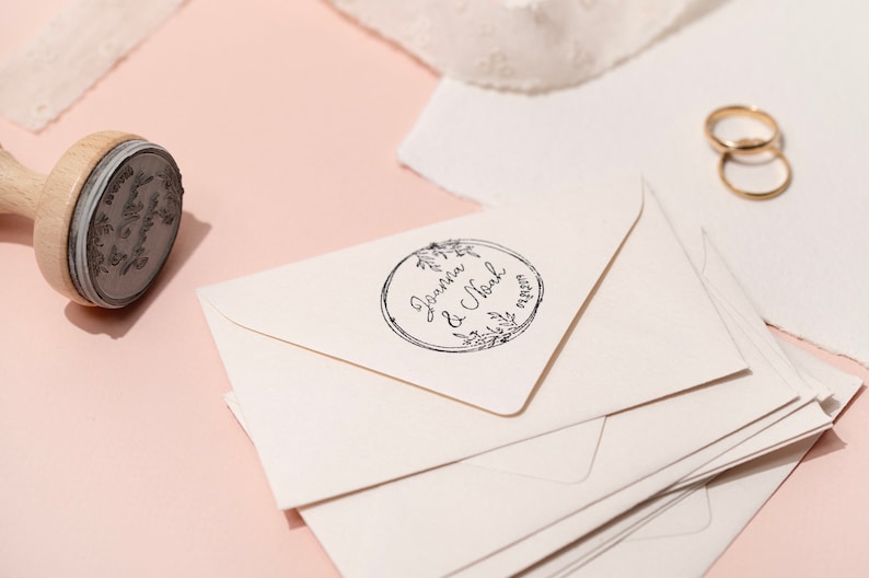 Op de afbeelding: Een houten stempel met een bloemenontwerp en de namen "Joanna & Noah" en een datum, naast een stapel cr&egrave;mekleurige enveloppen. Twee gouden trouwringen zijn zichtbaar op de achtergrond. De stempel staat op een lichtroze ondergrond.