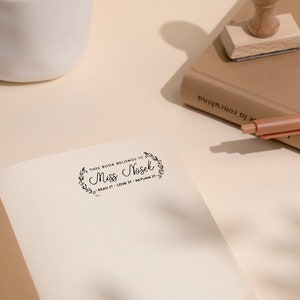 May include: An open book with a stamped inscription that reads "THIS BOOK BELONGS TO Miss Nosek READ IT - LOVE IT - RETURN IT." A wooden stamp and a pen rest on a closed book. A white ceramic vase is in the background.