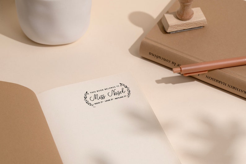 May include: An open book with a stamped inscription that reads "THIS BOOK BELONGS TO Miss Nosek READ IT - LOVE IT - RETURN IT." A wooden stamp and a pen rest on a closed book. A white ceramic vase is in the background.