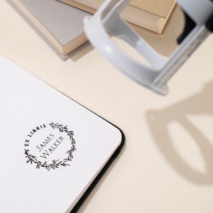 May include: A white notebook with a black border is stamped with a black "Ex Libris" design, including the name "James Walker" within a floral wreath. A gray stamp is positioned above the notebook, with a stack of books in the background.