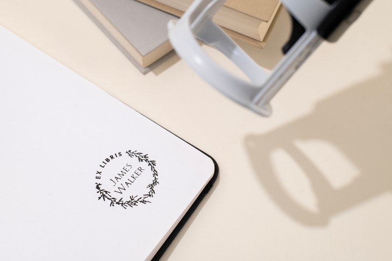 May include: A white notebook with a black border is stamped with a black "Ex Libris" design, including the name "James Walker" within a floral wreath. A gray stamp is positioned above the notebook, with a stack of books in the background.