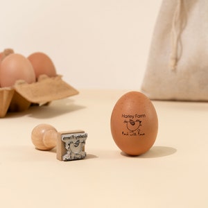 Puede incluir: Un huevo marr&oacute;n con un sello de goma que dice "Harley Farm" y "laid with love" con una peque&ntilde;a ilustraci&oacute;n de una gallina. Un sello de madera con el mismo dise&ntilde;o est&aacute; al lado del huevo.