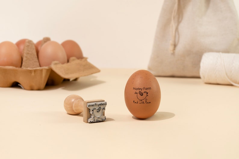 Puede incluir: Un huevo marr&oacute;n con un sello de goma que dice "Harley Farm" y "laid with love" con una peque&ntilde;a ilustraci&oacute;n de una gallina. Un sello de madera con el mismo dise&ntilde;o est&aacute; al lado del huevo.