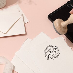 Op de afbeelding: Een houten stempel met de tekst "Save the Date" en een bloemenkransontwerp wordt gebruikt op een stapel getextureerd wit papier. Enveloppen en een klosje touw zijn ook zichtbaar op een roze oppervlak.