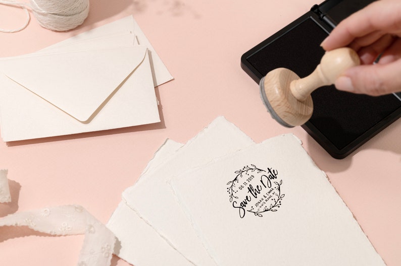 Op de afbeelding: Een houten stempel met de tekst "Save the Date" en een bloemenkransontwerp wordt gebruikt op een stapel getextureerd wit papier. Enveloppen en een klosje touw zijn ook zichtbaar op een roze oppervlak.