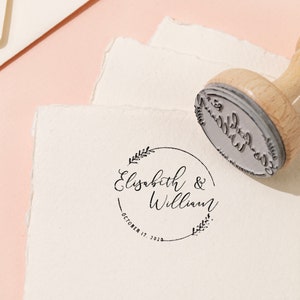 Op de afbeelding: Een houten stempel met een cirkelvormig ontwerp, met de namen "Elisabeth & William" en de datum "October 17, 2023". De stempel staat op een wit papier met een gescheurde rand, met gouden ringen en briefpapier op de achtergrond.