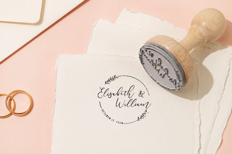 Op de afbeelding: Een houten stempel met een cirkelvormig ontwerp, met de namen "Elisabeth & William" en de datum "October 17, 2023". De stempel staat op een wit papier met een gescheurde rand, met gouden ringen en briefpapier op de achtergrond.