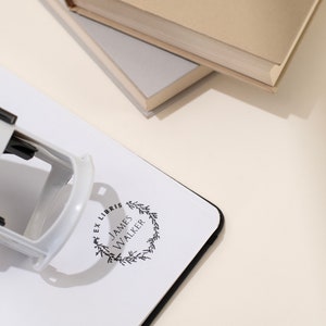 May include: A black and white self-inking stamp with the text "EX LIBRIS JAMES WALKER" inside a wreath design. The stamp is on a white surface with a black border, near two books.