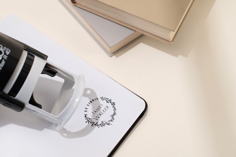 May include: A black and white self-inking stamp with the text "EX LIBRIS JAMES WALKER" inside a wreath design. The stamp is on a white surface with a black border, near two books.