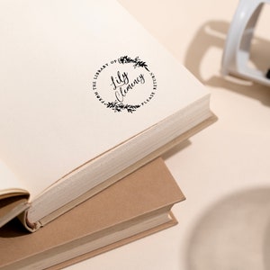 May include: A close-up of a book with a round, black stamp that reads "From the Library of Lily Clemency Please Return". The stamp is surrounded by a wreath of leaves.