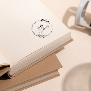 May include: An open book with a stamped design that reads "From the Library of Lily Clemency, Please Return" in a circular floral wreath. A white and black stamp is in the background.