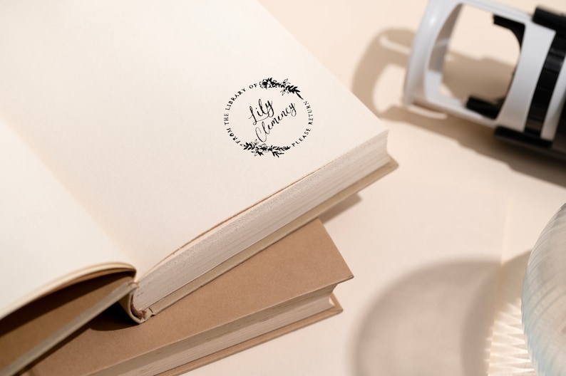 May include: An open book with a stamped design that reads "From the Library of Lily Clemency, Please Return" in a circular floral wreath. A white and black stamp is in the background.