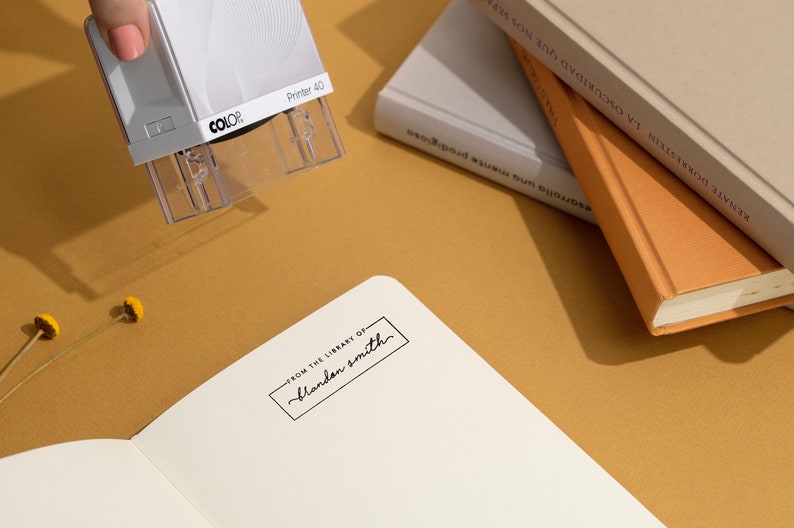 May include: A white self-inking stamp with the text "COLOR Printer 40" on it. The stamp is being used to stamp a book with the text "FROM THE LIBRARY OF blandan smith".