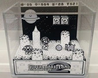 May include: A black and white pixel art diorama of a scene from Super Mario Land 2: 6 Golden Coins. The diorama features Mario in a space suit, a Goomba, a Koopa Troopa, and a question block.