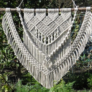 Könnte beinhalten: Ein großer, cremefarbener Makramee-Wandbehang mit geometrischem Muster und Fransen. Der Behang ist aus natürlicher Baumwollschnur gefertigt und ein beliebtes Dekoelement im Boho-Stil.