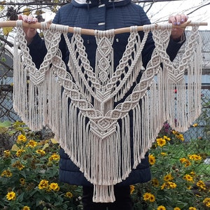 Könnte beinhalten: Ein großer, beiger Makramee-Wandbehang mit geometrischem Muster und Fransen. Der Wandbehang ist aus natürlicher Baumwollschnur gefertigt und an einer Holzleiste befestigt.
