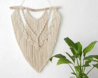 Puede incluir: Un tapiz de macramé beige con una rama de madera en la parte superior. El tapiz presenta un patrón geométrico con detalles de flecos.