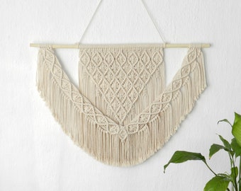 May include: A beige macrame wall hanging with a geometric pattern and fringe. The hanging is attached to a wooden dowel rod.