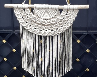 May include: A white macrame wall hanging with a wooden dowel at the top and a fringe at the bottom. The macrame is in a geometric pattern with a series of arches.
