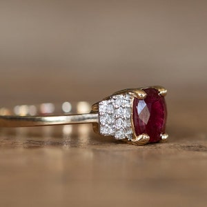 Puede incluir: Un anillo de oro con un gran rubí ovalado. El rubí es de color rojo intenso y está acentuado por piedras preciosas más pequeñas y claras. El anillo está engastado en una banda de oro, mostrando un diseño clásico y elegante.