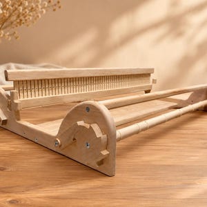 May include: A light-colored wooden weaving loom featuring a rigid heddle with vertical slots, two side support panels with gear-like adjustment wheels, and two horizontal cylindrical rods for tensioning and holding warp threads in place.