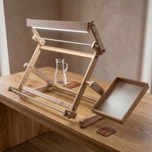 May include: A light-colored wooden embroidery frame stand featuring an adjustable frame with a built-in LED light bar, metal tension rods, and a side-mounted wooden tray. The structure includes wing nuts for angle adjustments and a notched base.