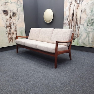 Puede incluir: Sofá de tres plazas de estilo moderno de mediados de siglo, con tela clara y estructura de madera. El sofá tiene un diseño minimalista con líneas limpias. La habitación presenta arte abstracto y una paleta de colores neutros.