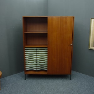 Pode incluir: Um armário de madeira de estilo moderno de meados do século com uma porta deslizante e prateleiras. O armário tem um acabamento de madeira clara e está sobre pernas de metal. O armário tem um organizador de arquivos integrado com pastas brancas.