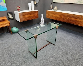Puede incluir: Una mesa consola rectangular con una superficie de cristal transparente y dos paneles laterales verticales de cristal. Los componentes de cristal están unidos por herrajes de metal color bronce y la base se apoya en un marco metálico fino y oscuro.