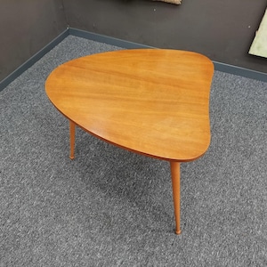 Könnte beinhalten: Ein Couchtisch im Mid-Century-Modern-Stil mit drei Beinen. Die Tischplatte hat eine hellbraune, organische Form. Die Beine sind in einem passenden Hellbraun gehalten. Der Tisch ist etwa 76 cm lang.