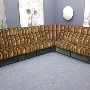 May include: A large, sectional sofa with a curved design. The sofa features vertical stripes in shades of brown, orange, and green. The base and armrests are a solid dark green. The sofa is placed in a room with artwork on the walls.