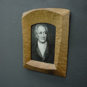 Peut inclure: Un portrait en noir et blanc d'un homme dans un cadre en bois sculpté. L'homme porte une chemise blanche avec une veste noire et a une expression sérieuse sur son visage.