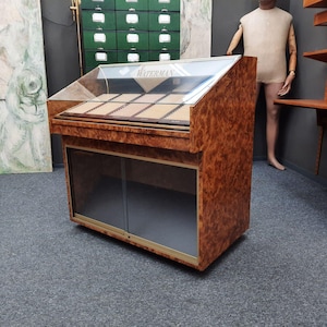 Puede incluir: Una gramola de madera vintage con tapa de cristal y puertas correderas de cristal. La gramola tiene un cálido acabado de madera de raíz y presenta el texto "Waterman" en la parte superior. Es un mueble clásico.